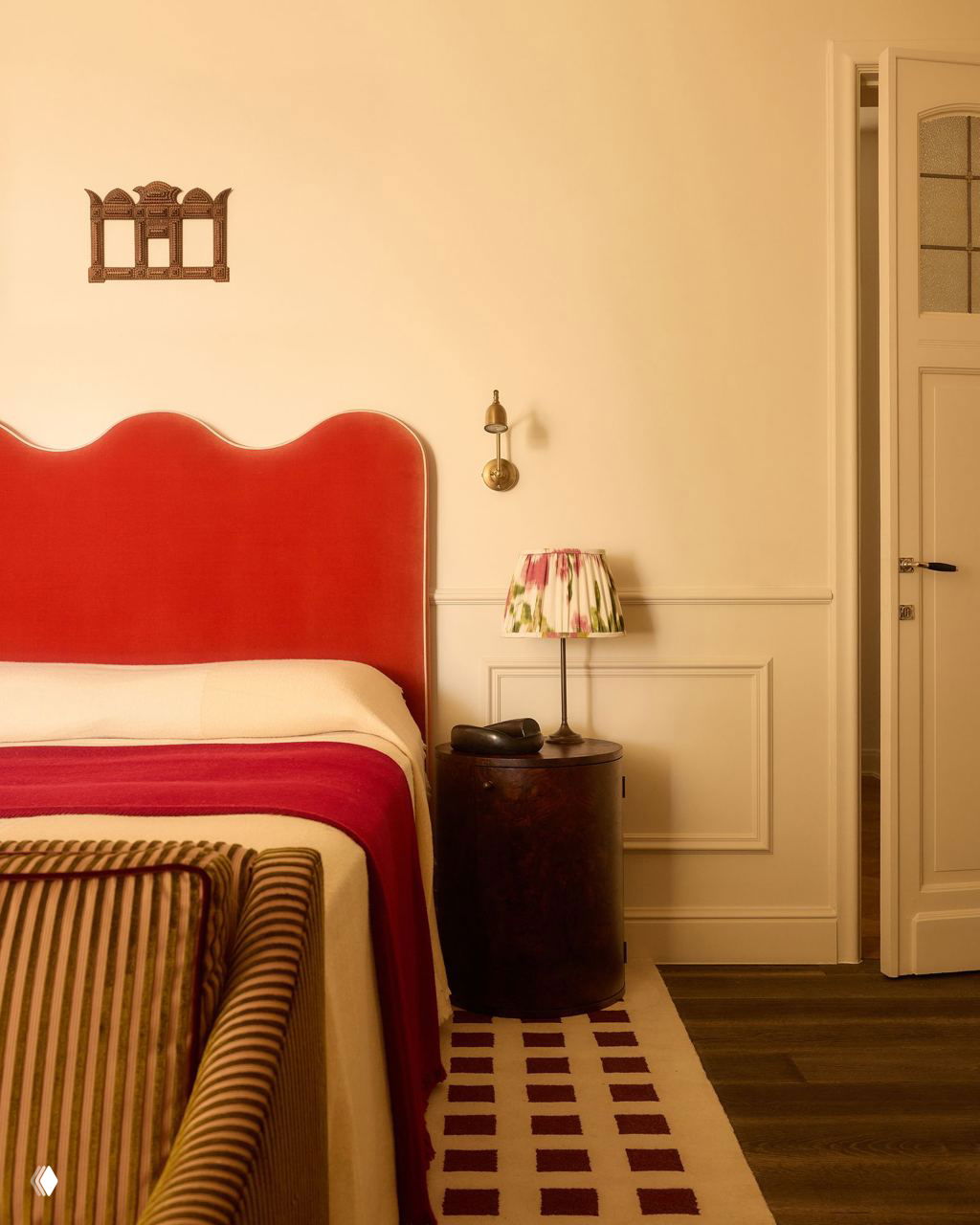 Спальня с ярко‑красным изголовьем кровати, спокойными текстурами постельного белья и ковром с геометрическим рисунком на нейтральном фоне стен.