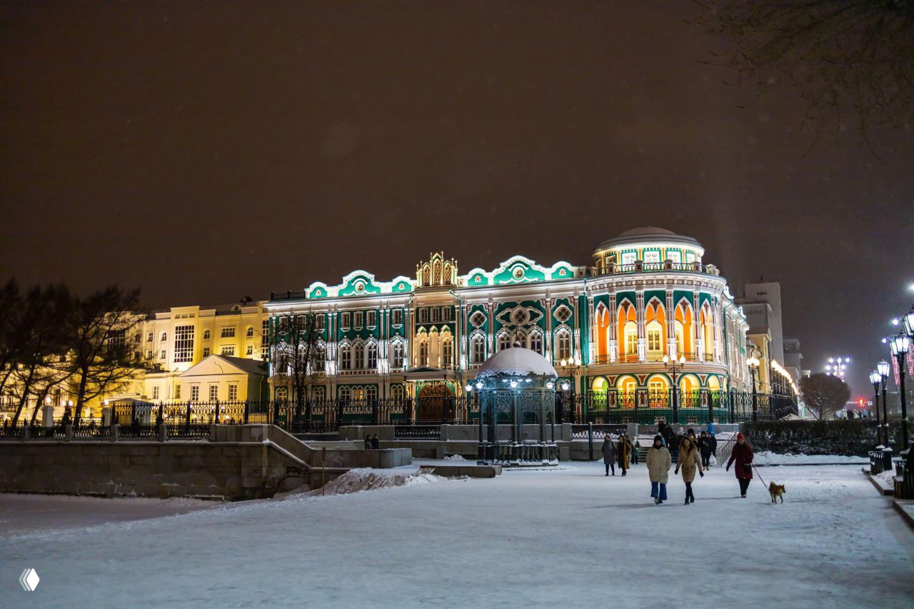 Дом Севастьянова вечером в праздничной подсветке, заснеженная набережная и прохожие, уютные огни и архитектурные детали фасада на фоне неба.
