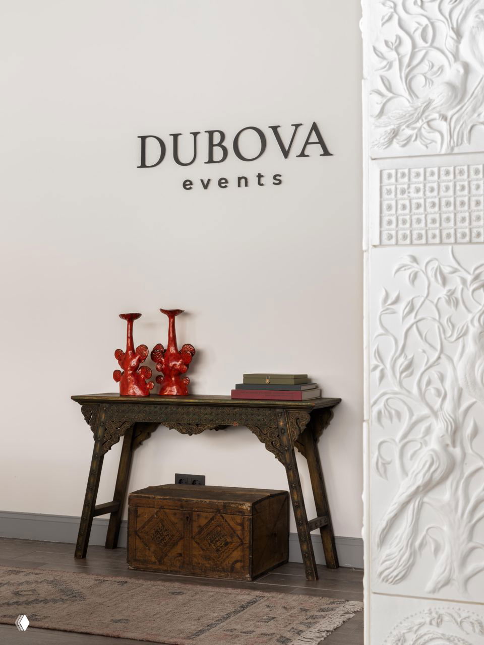 Деревянная консоль у стены с логотипом DUBOVA events, старинный сундук под столом и декоративные красные сувенирные предметы на поверхности.