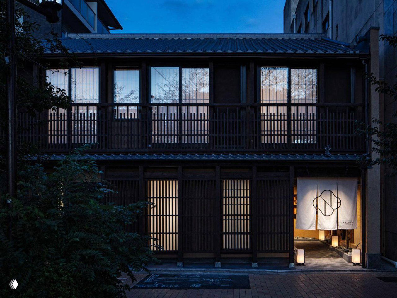 Ночной фасад переработанного столетнего матия в Киото с традиционной решёткой, занавесом у входа и приглушённым светом от отеля Nazuna Kyoto Higashihonganji