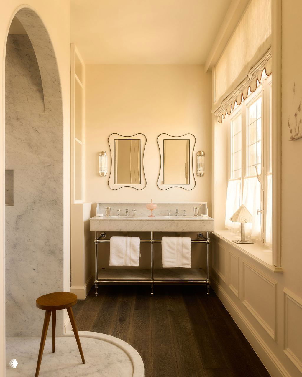 Ванная комната с двойной мраморной раковиной, двумя зеркалами в фигурных рамах и тёмным деревянным полом, свет льётся через большое окно.