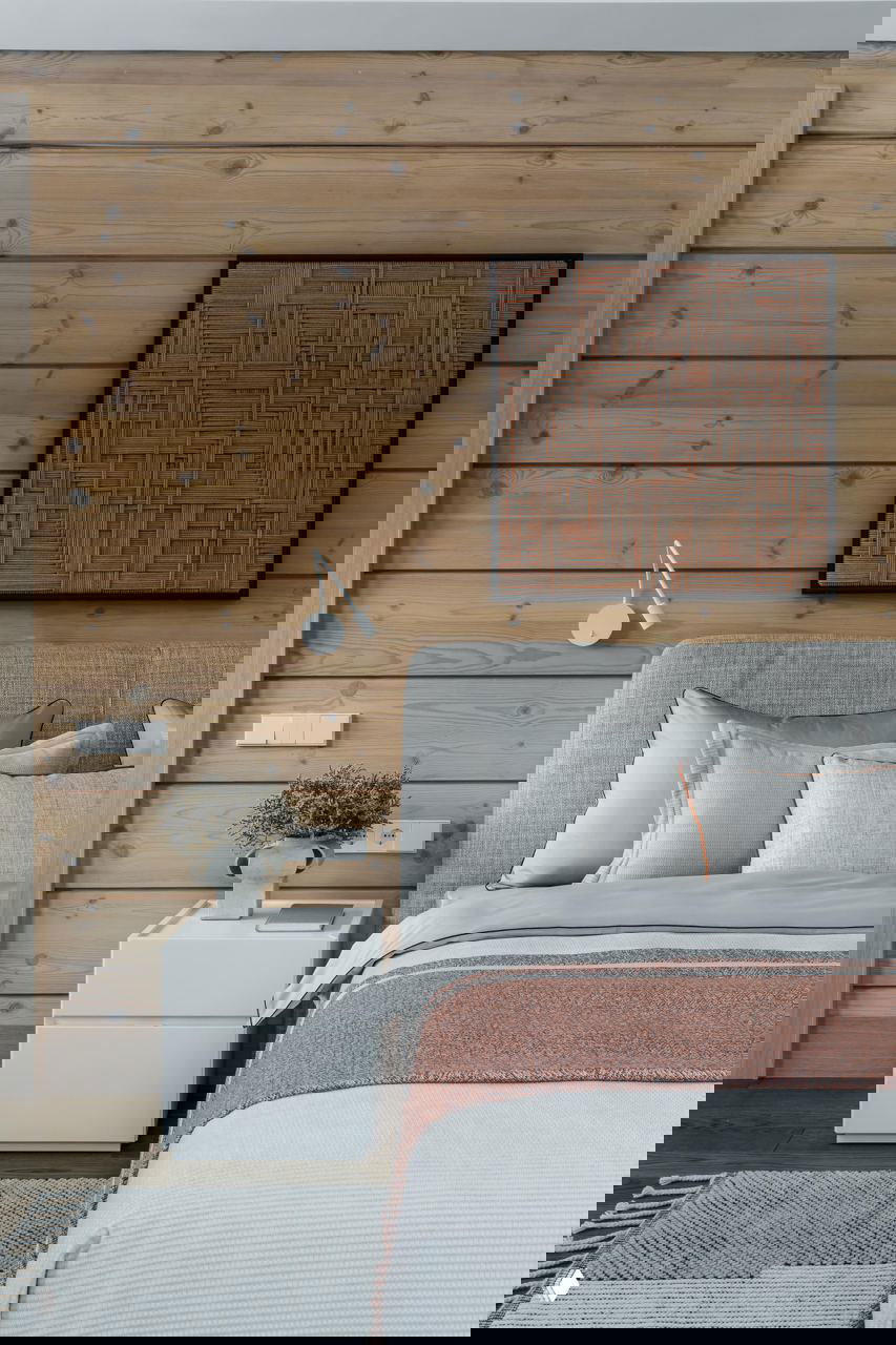 Спальня с деревянной стеной из клееного бруса, мягким изголовьем и текстильными слоями в спокойных серых и бежевых тонах, акцентный текстиль.