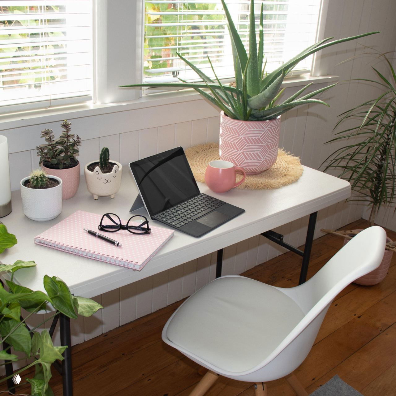 Рабочий стол у окна с планшетом, очками, блокнотом, кружкой и несколькими горшочными растениями, среди которых маленькие кактусы в декоративных горшках, уютный интерьер.