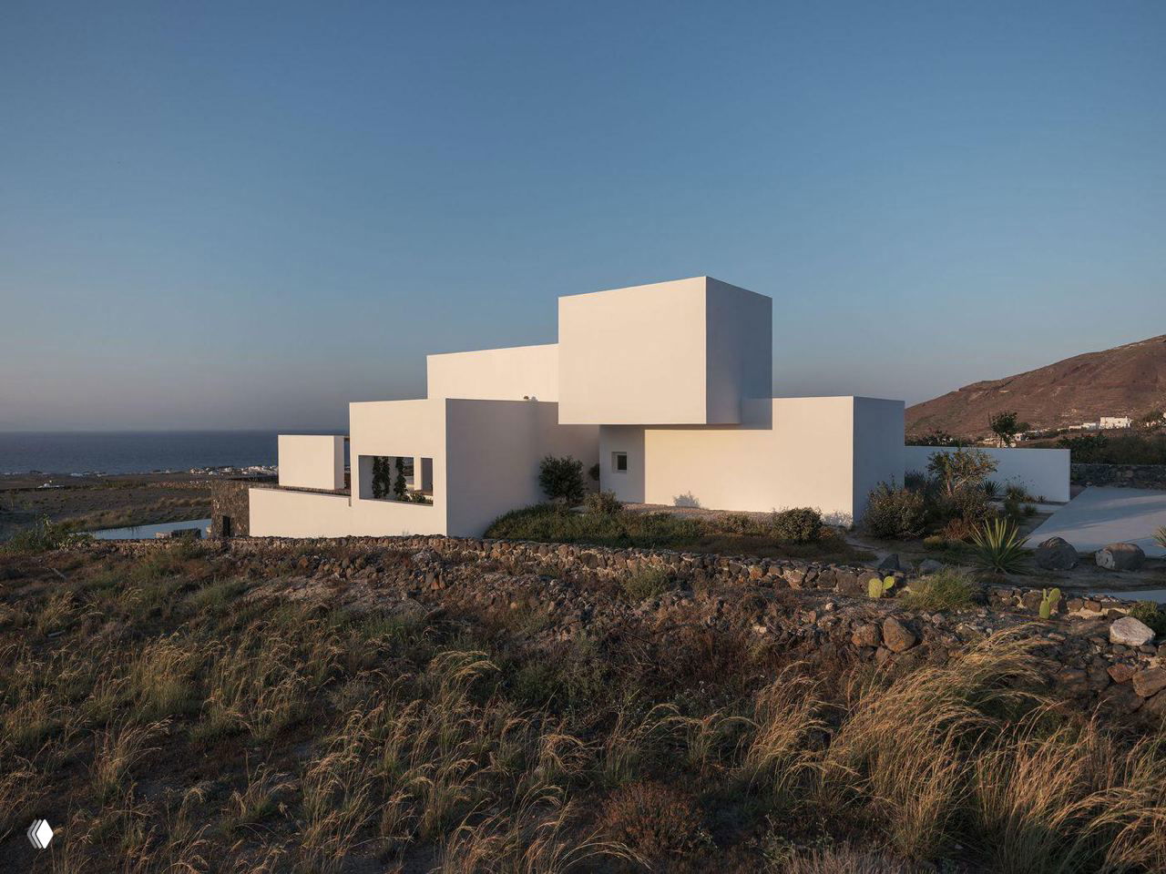 Экстерьер виллы на Санторини: белые кубические объёмы Kapsimalis Architects на фоне моря и каменистой прибрежной растительности.
