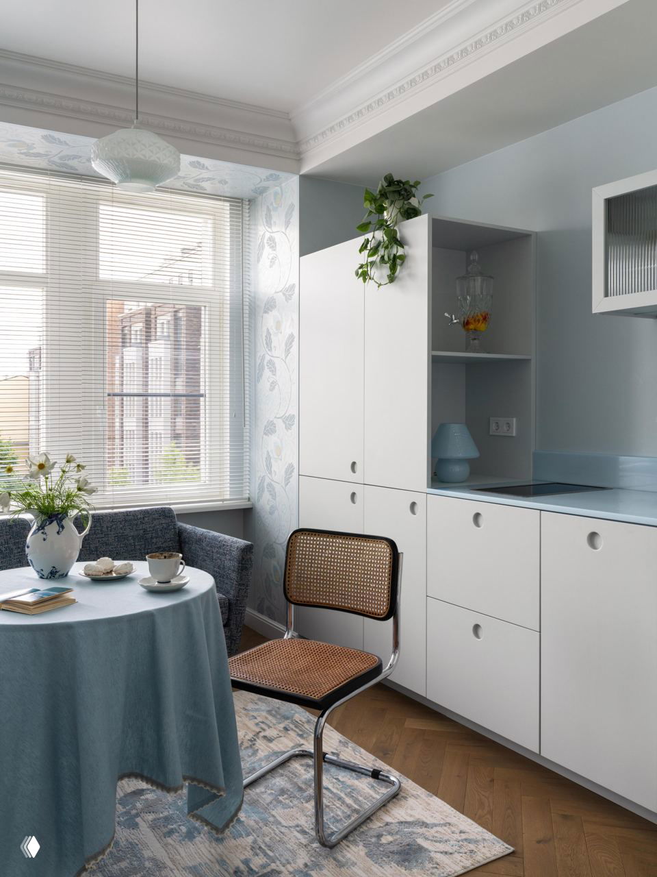 Кухонная зона в пастельных голубых тонах: круглый стол со скатертью, ротанговый стул, белые встроенные шкафы, окно с жалюзи и горшечное растение.