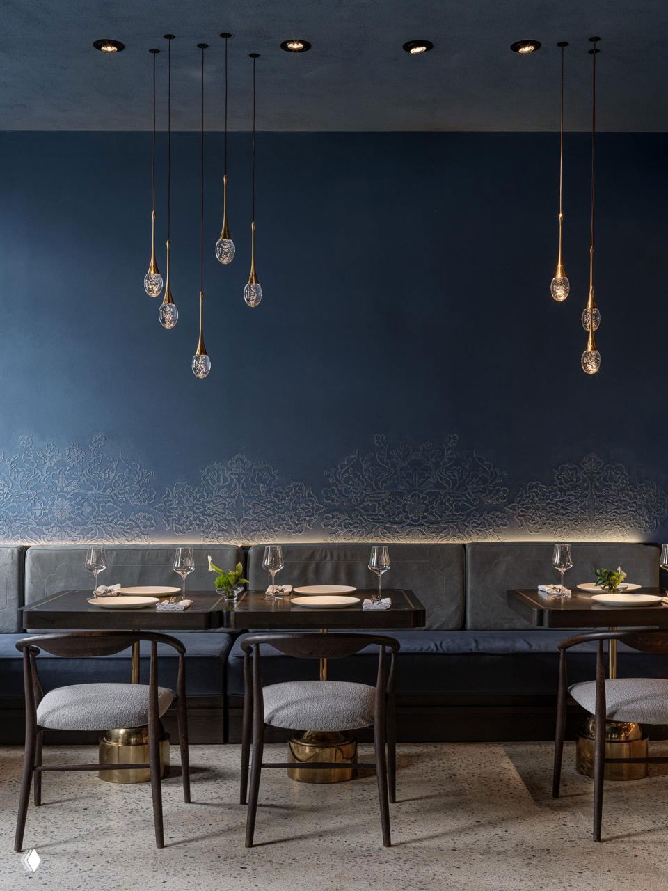 Ряд посадочных мест вдоль стены: низкие диваны, деревянные столы и декоративный рельефный орнамент на тёмно‑синей фактурной стене, над ними каплевидные светильники.