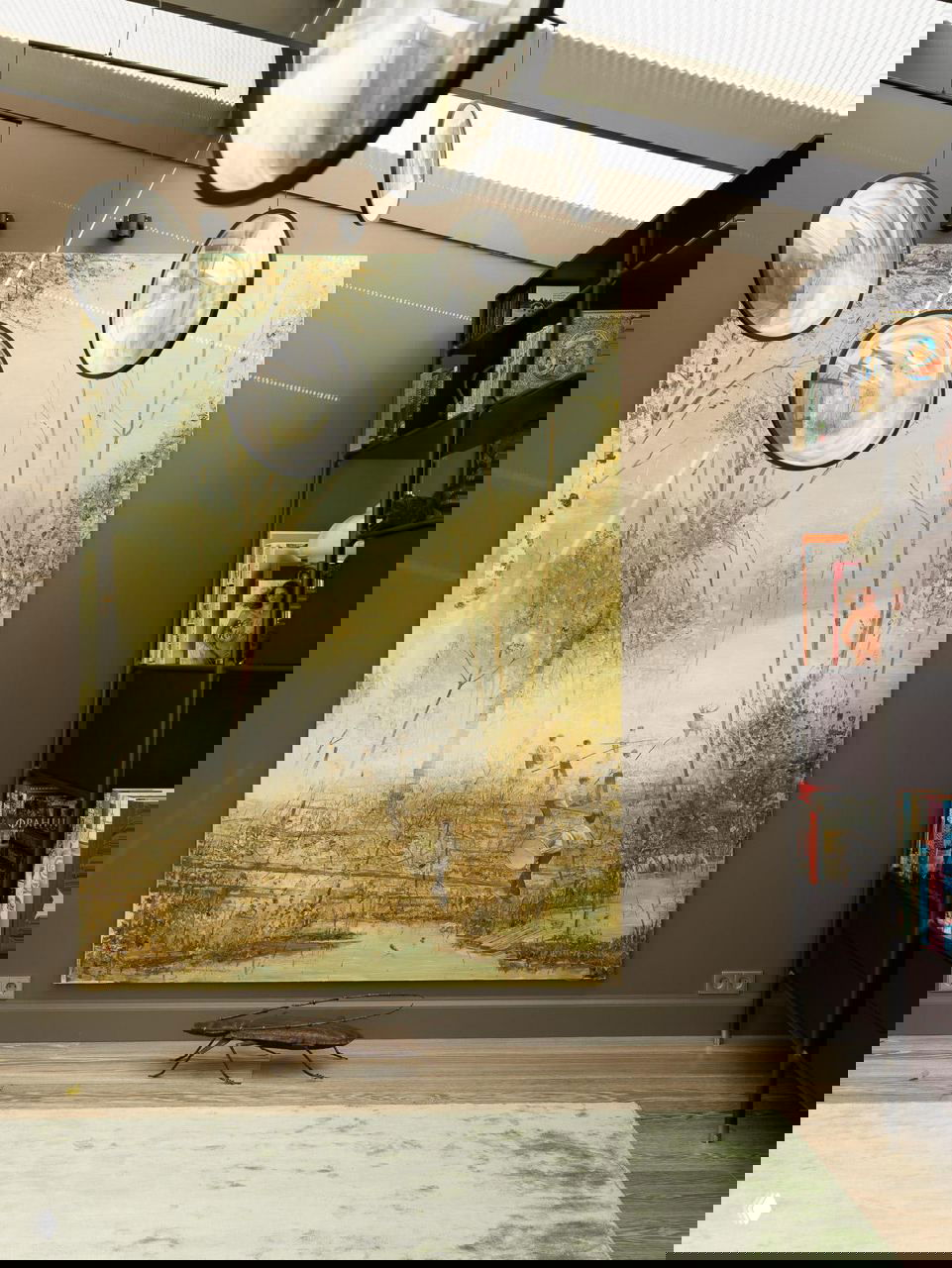 Высокое настенное живописное панно пасторальной сцены в светлых тонах; рядом книжный стеллаж и подвесные круглые элементы декора под потолком.