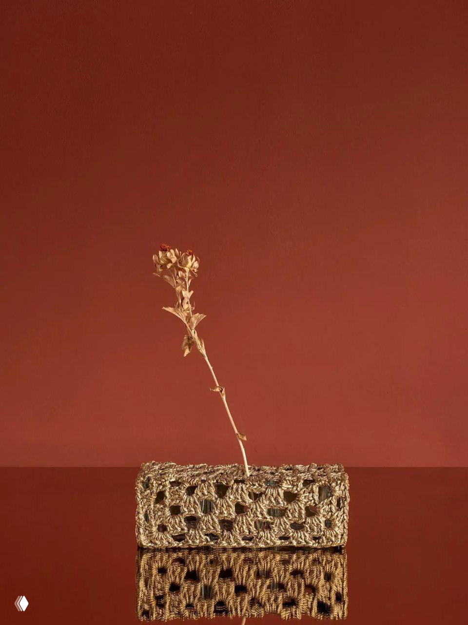 Небольшой предмет, выглядящий как вязаная сумочка, отлитая в бронзе, на фоне тёмно-красной стены с одиночной сухой веткой — предмет от Nilufar Gallery.