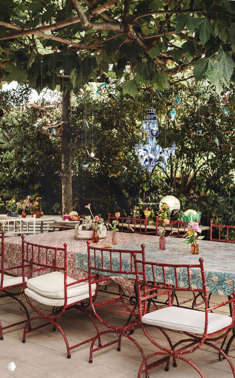 Открытая столовая под деревьями с длинным столом, разноцветными огнями и плетёными стульями — неформальная внешняя обеденная зона в саду.