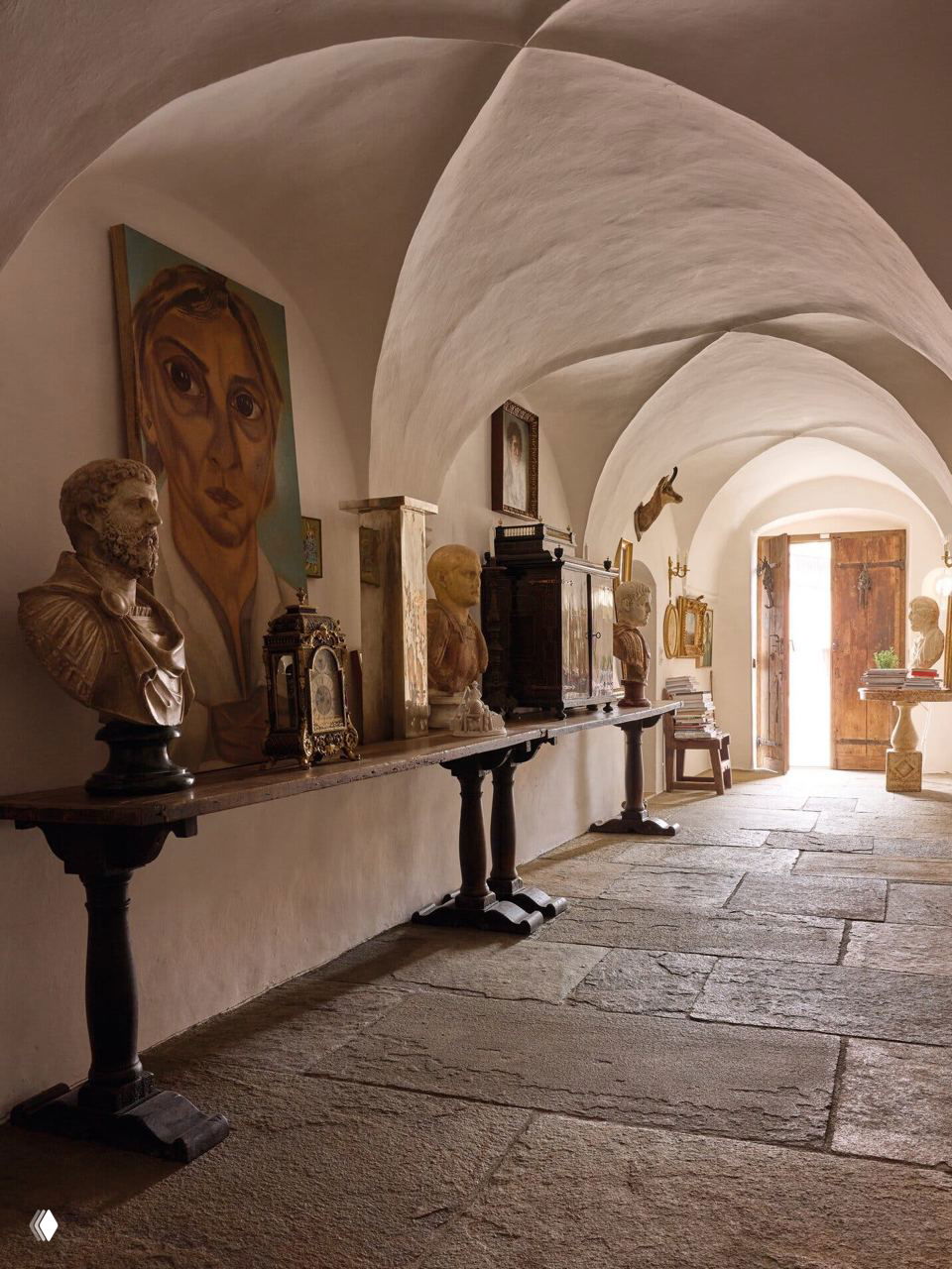 Длинный арочный коридор поместья Джана Энцо Спероне: грубый каменный пол, стол с бюстами, картинами и старинными предметами, свет у открытой двери.