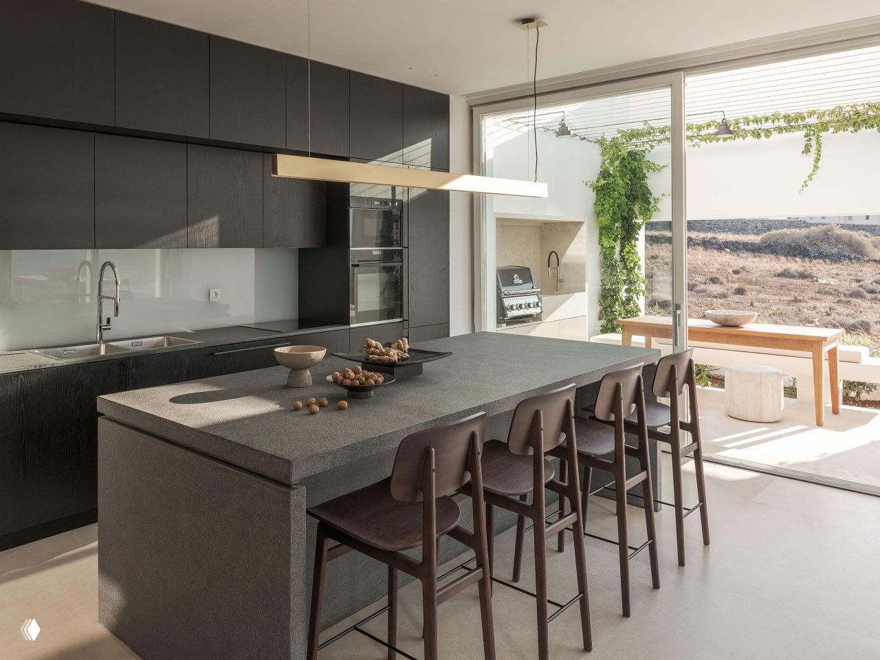 Кухонная зона внутри виллы: строгие тёмные шкафы, остров и барные стулья на фоне открытого выхода к террасе и гриль-зоне.