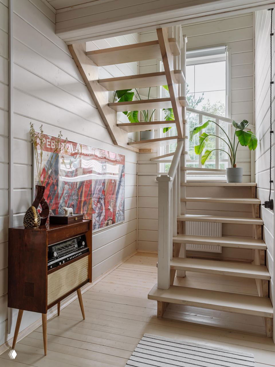 Световая лестница с открытыми ступенями, растения у окна и винтажный радиоприёмник на тумбе — переходная зона с характером дачного дома.