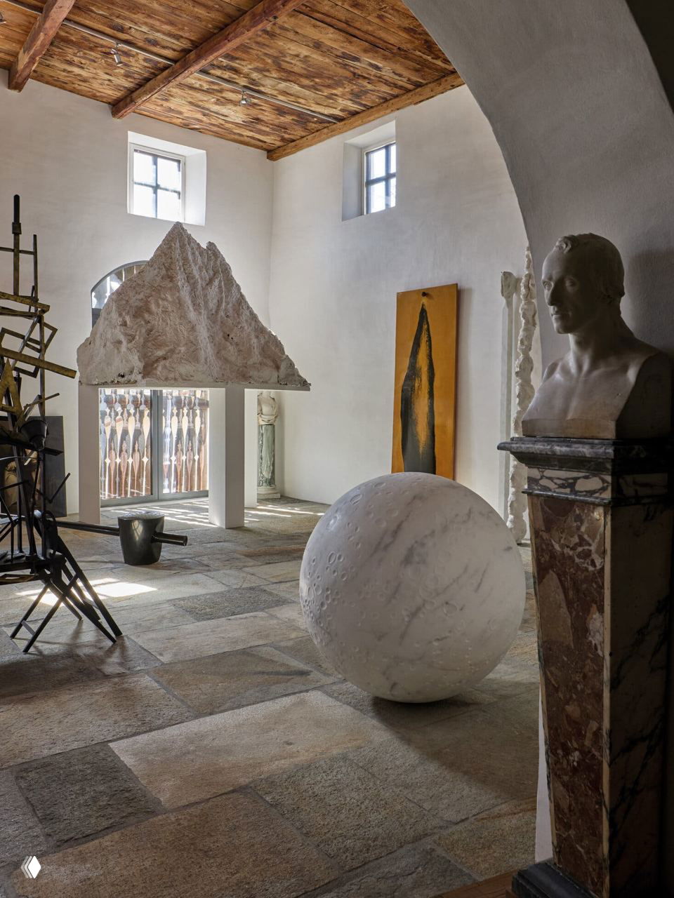 Светлая галерея в поместье Спероне с крупными современными скульптурами — мраморный шар, рельеф и металличесные объекты на фоне высоких окон и деревянных балок.
