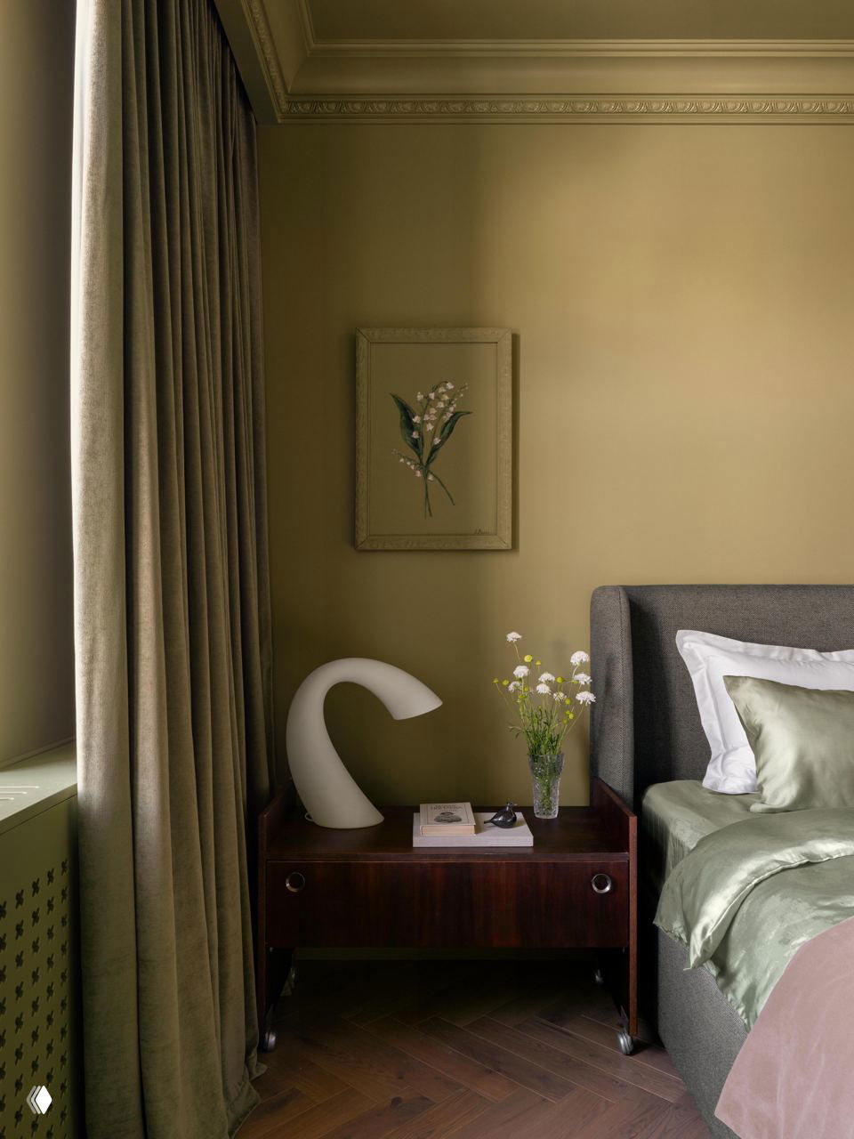 Прикроватная зона: деревянная тумбочка тёмного дерева, современная лампа и ваза с полевыми цветами на фоне зелёной стены.