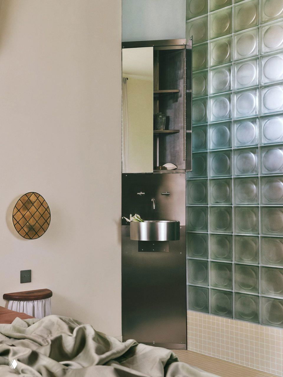 Угловой фрагмент спальни и ванной: стеклоблоки образуют приватную перегородку, на фоне видна металлическая раковина и деревянная отделка.