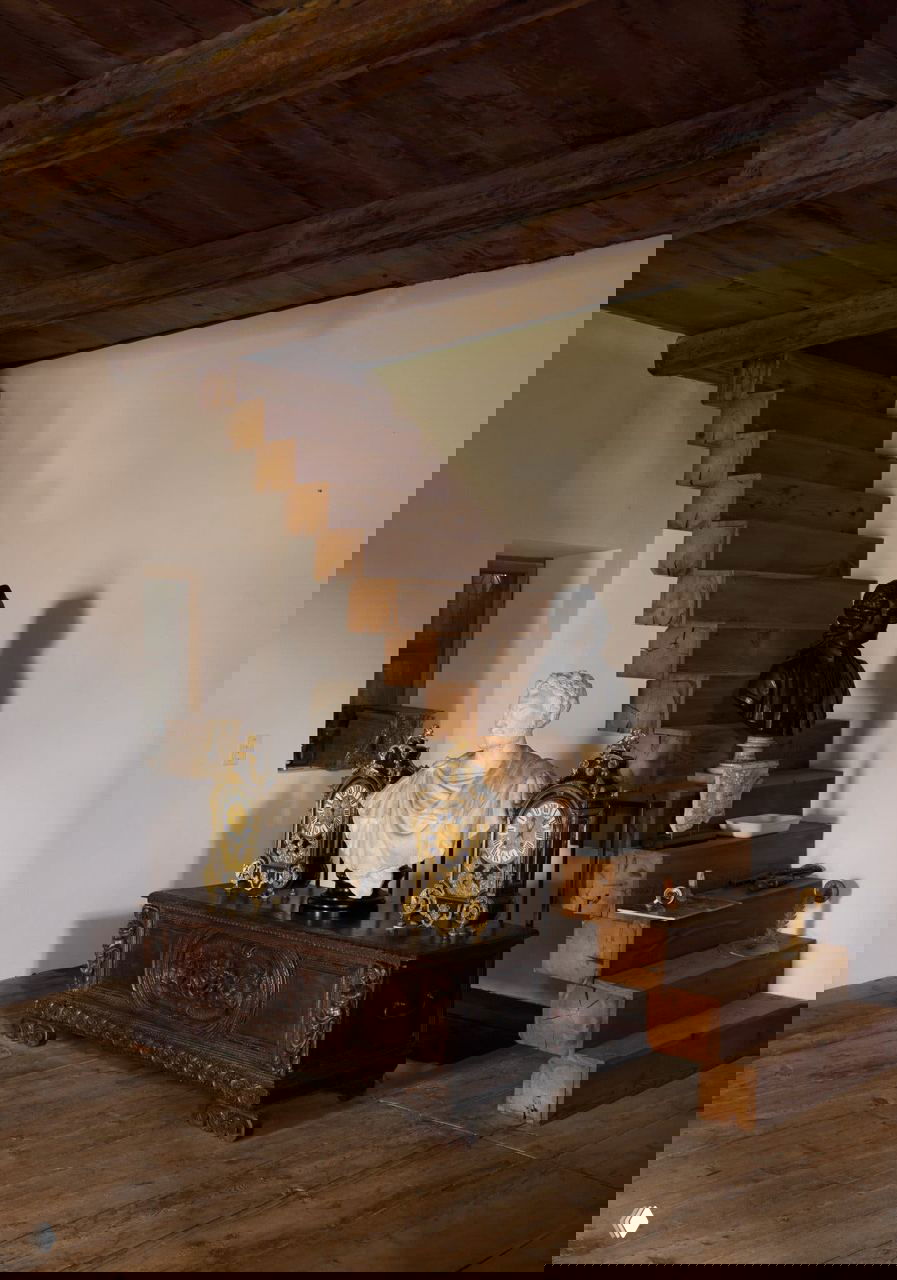 Пространство у деревянной лестницы в поместье: антикварный комод, мраморный бюст и пара старинных настольных часов на фоне массивных ступеней и тёплого пола.