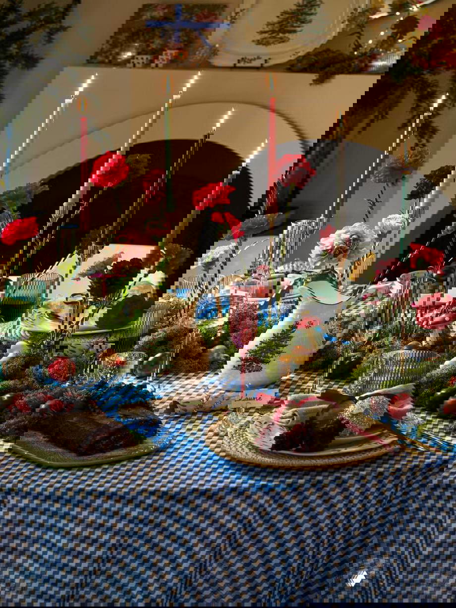 Праздничная сервировка на столе: высокие цветные свечи, свежие цветы и клетчатая скатерть; композиция в стиле домашнего праздничного обеда с декором.