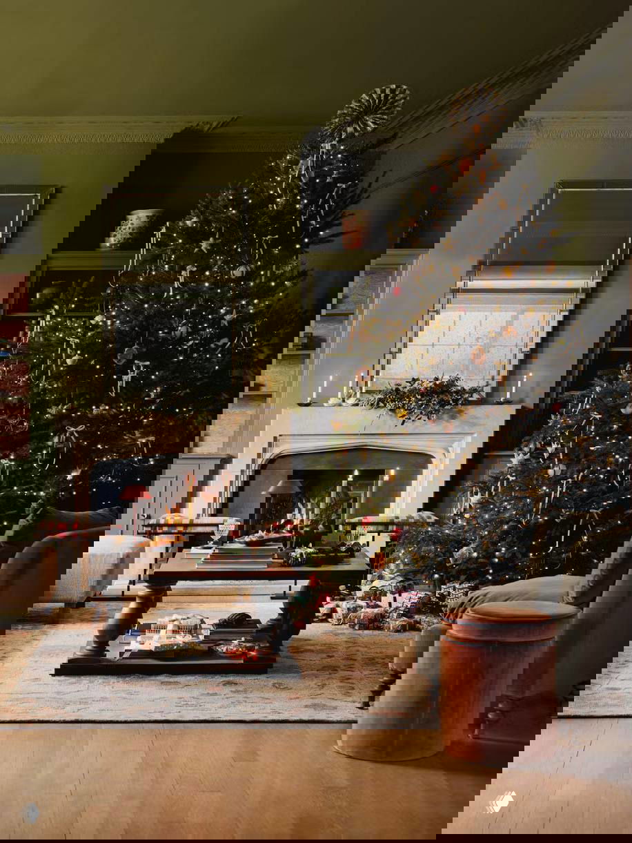 Уютная гостиная с тёмно-зелёной стеной и большой рождественской ёлкой у камина; подарки, свечи и тёплое мерцание гирлянд создают праздничную атмосферу.