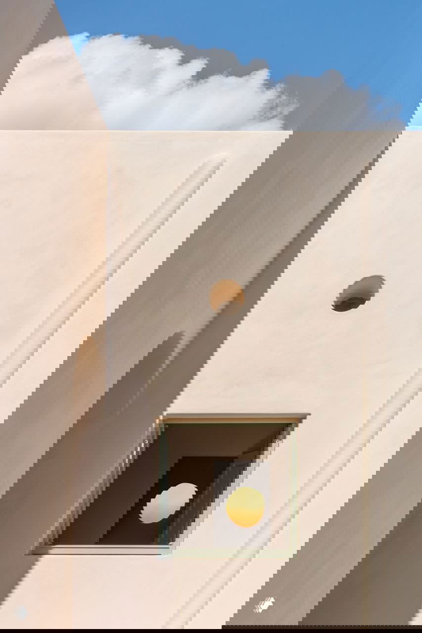 Фрагмент фасада с гладкими бежевыми стенами, квадратным окном и круглым отверстием, игра света и тени на поверхности стены.