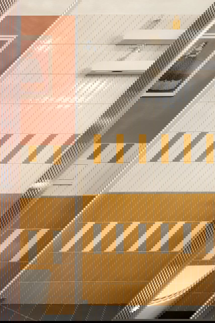 Узкая ванная с декоративной плиткой: белые вертикальные плитки сочетаются с графичной жёлто-терракотовой вставкой и чёрным полом.