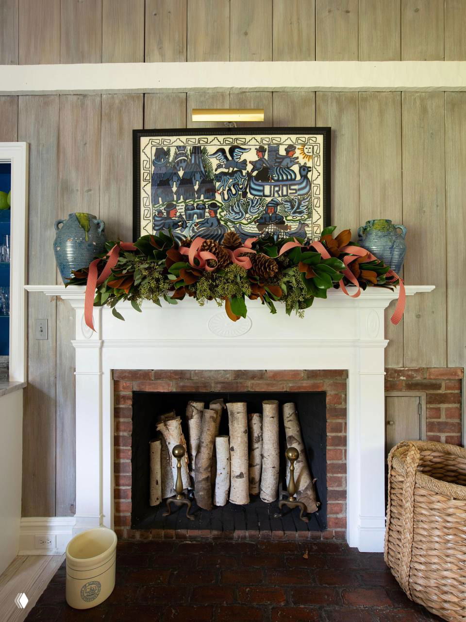 Камин с белой облицовкой украшен гирляндой из хвои и шишек; в топке декоративно уложены берёзовые поленья, интерьер в светлых тёплых тонах.