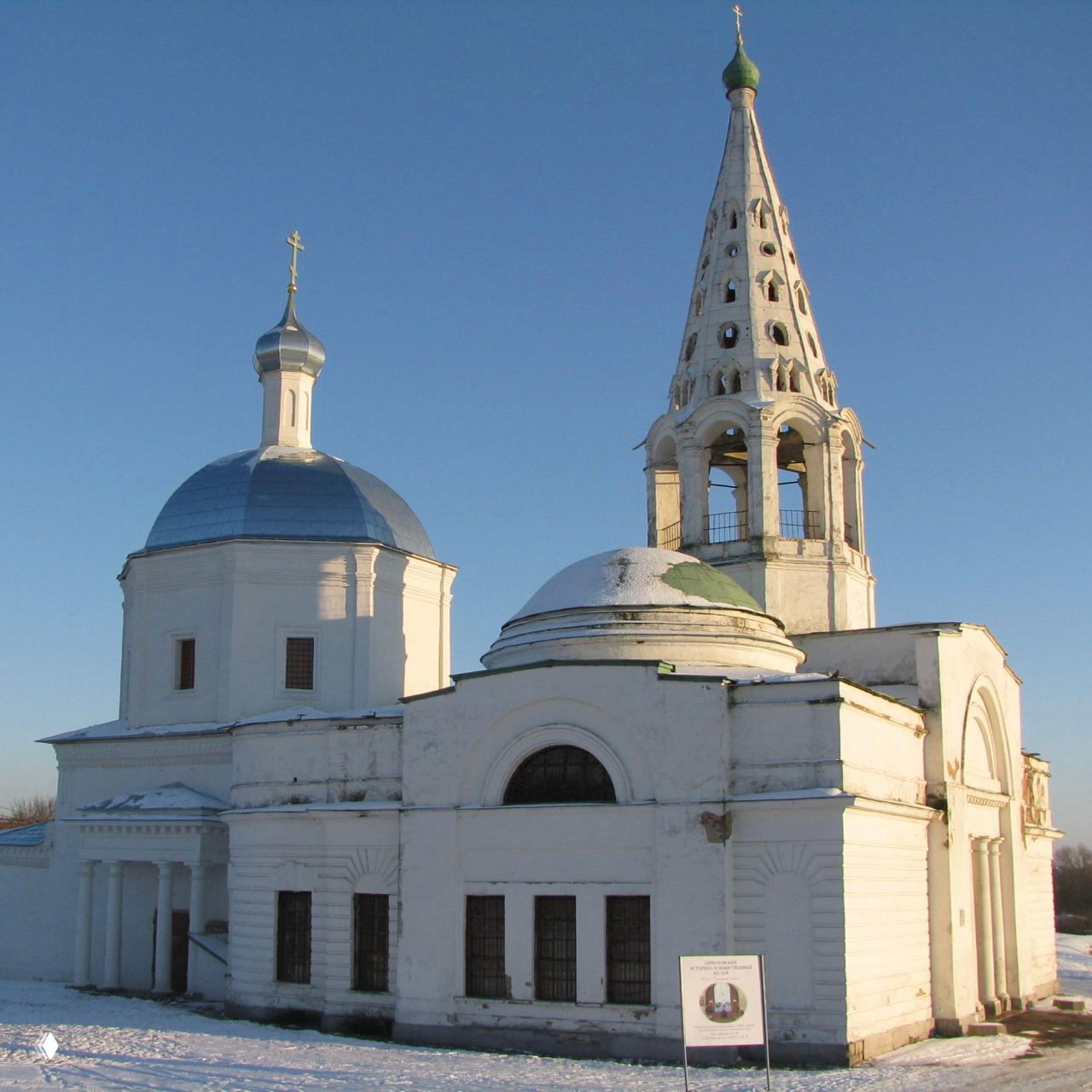 Белая церковь с куполом и шатровой звонницей на фоне ясного голубого неба и равномерного снежного покрова вокруг здания.