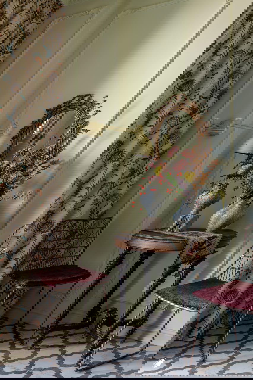 Невысокий круглый стол с вазами и зеркало в резной раме на фоне мягкой зелёной панели и цветочных штор в винтажном стиле.