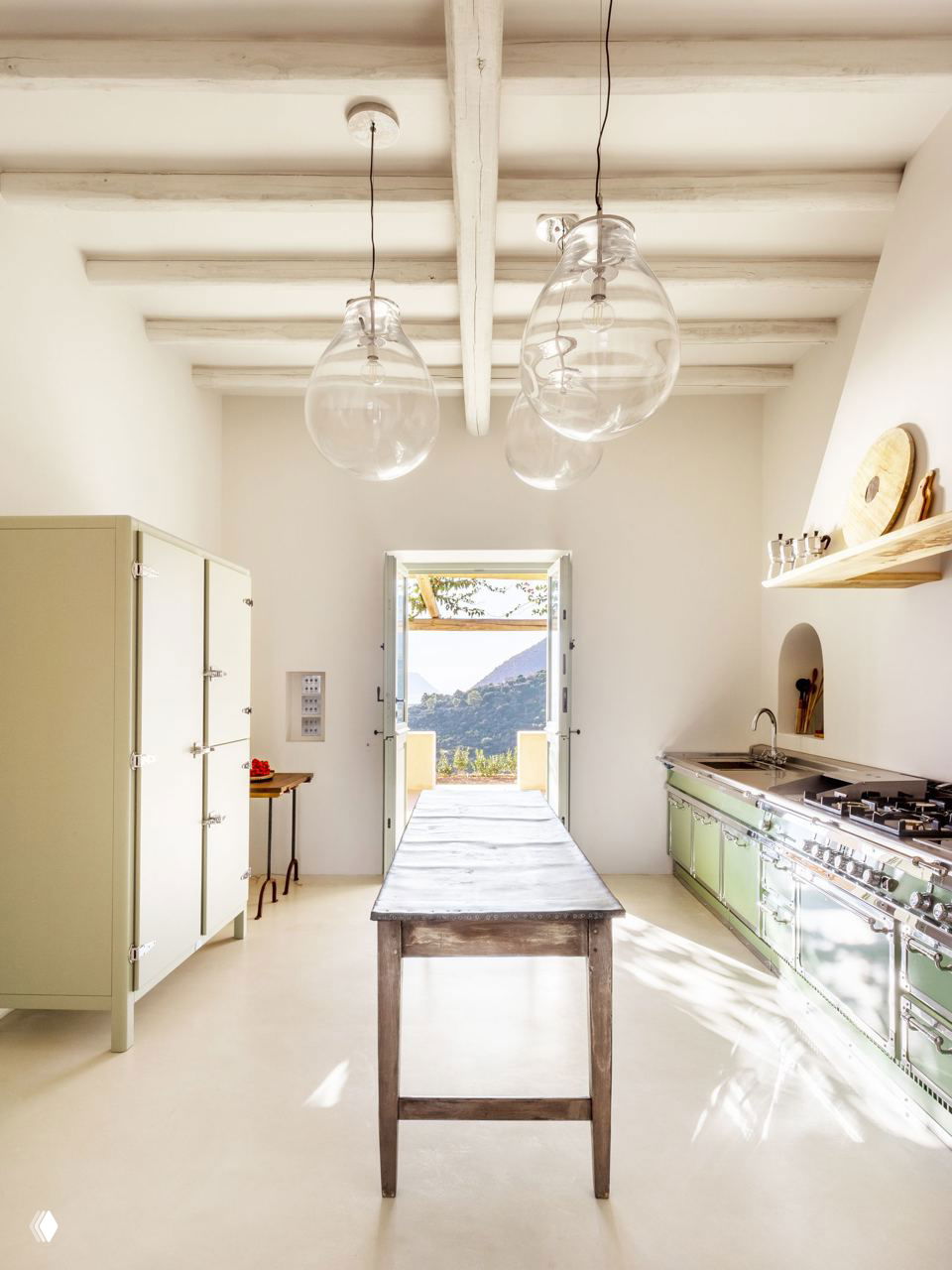 Кухонное пространство в светлых тонах: видна длинная столешница, винтажная зелёная кухня, деревянные балки и дверь на террасу с видом наружу.