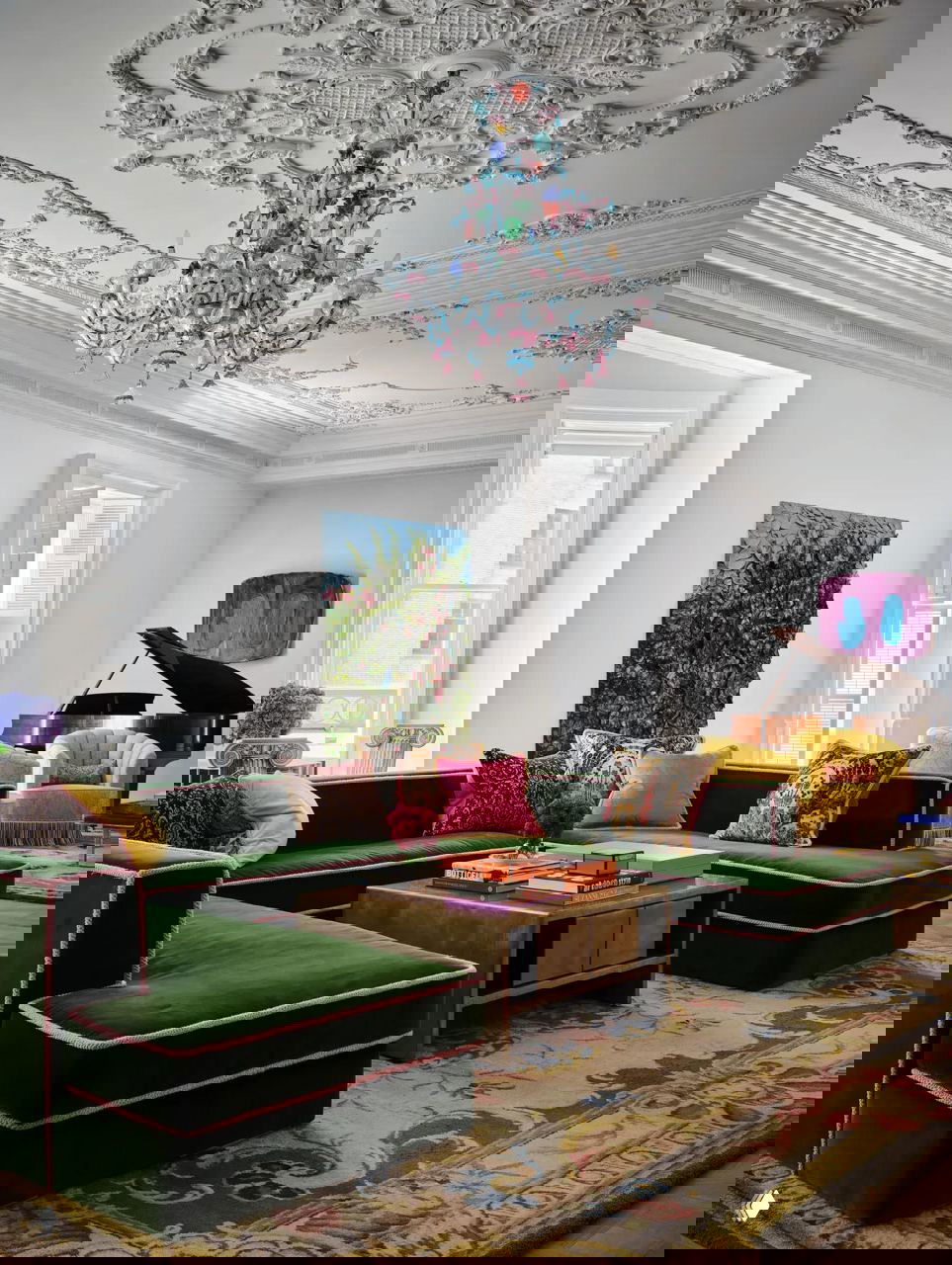 Гостиная с зелёным бархатным модульным диваном, цветной муранской люстрой, лепным белым потолком и большим роялем на заднем плане.