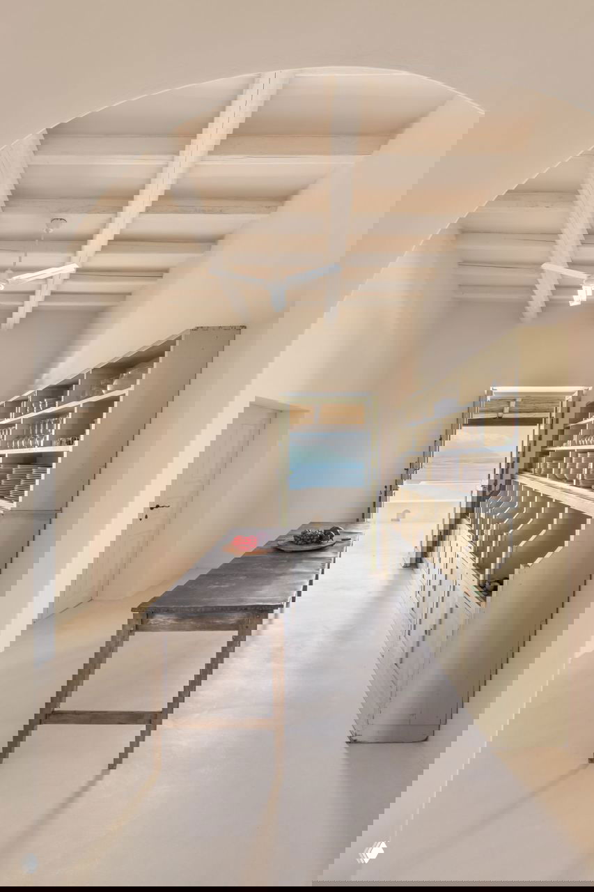 Проход с высокой аркой и длинным столом, аккуратные открытые полки и устремлённая к свету перспектива к выходу на террасу и морю.