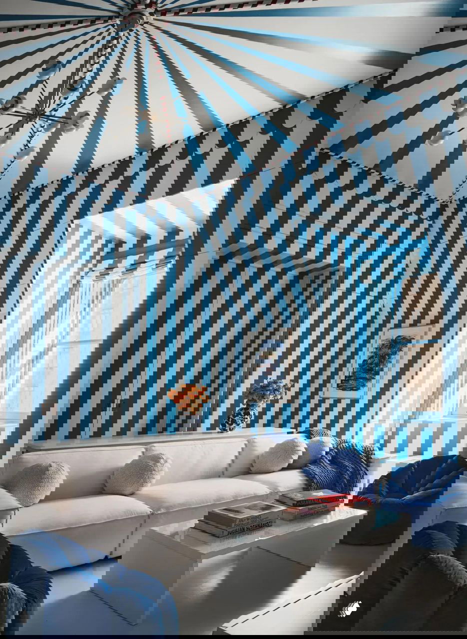 Комната с белым диваном на фоне стен и потолка в синие вертикальные полосы, низкая белая кофейная столешница и синие пуфы.