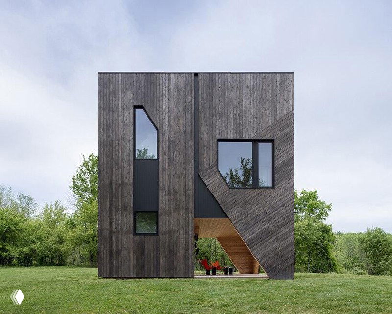 Крупный ракурс фасада коттеджа под углом: темная облицовка и окна, вокруг — зеленая лужайка; на фото заметны цифровые артефакты.