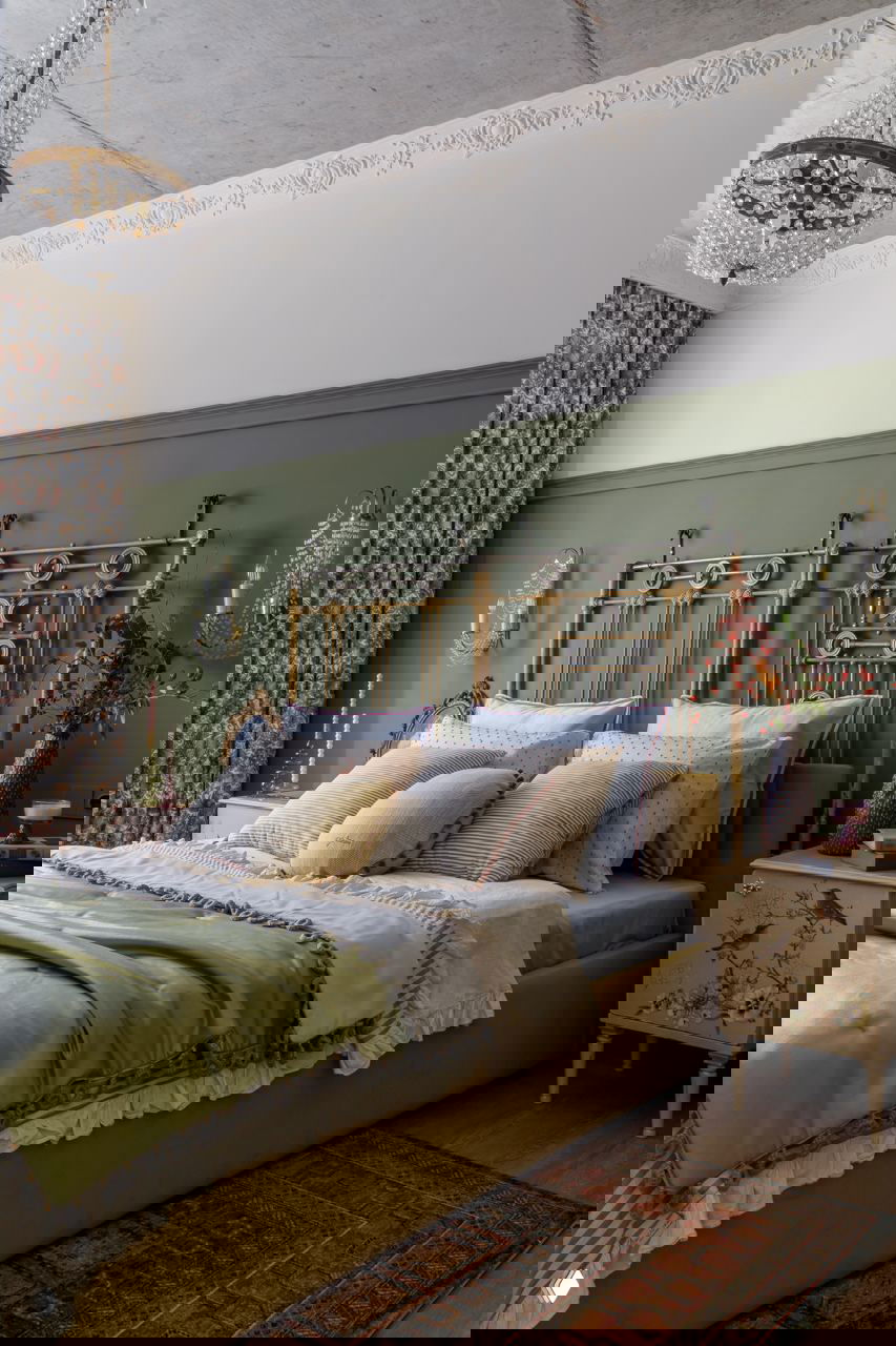 Спальня с металлическим винтажным изголовьем, зелёной стеной под панелью, текстильными подушками и кристаллической люстрой на контрастном потолке.