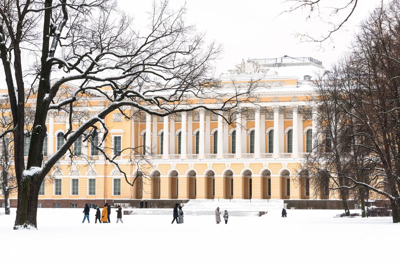 Заснеженный городской парк и фасад дворцового здания в Санкт-Петербурге зимой, редкие прохожие на белоснежной лужайке перед колоннами.