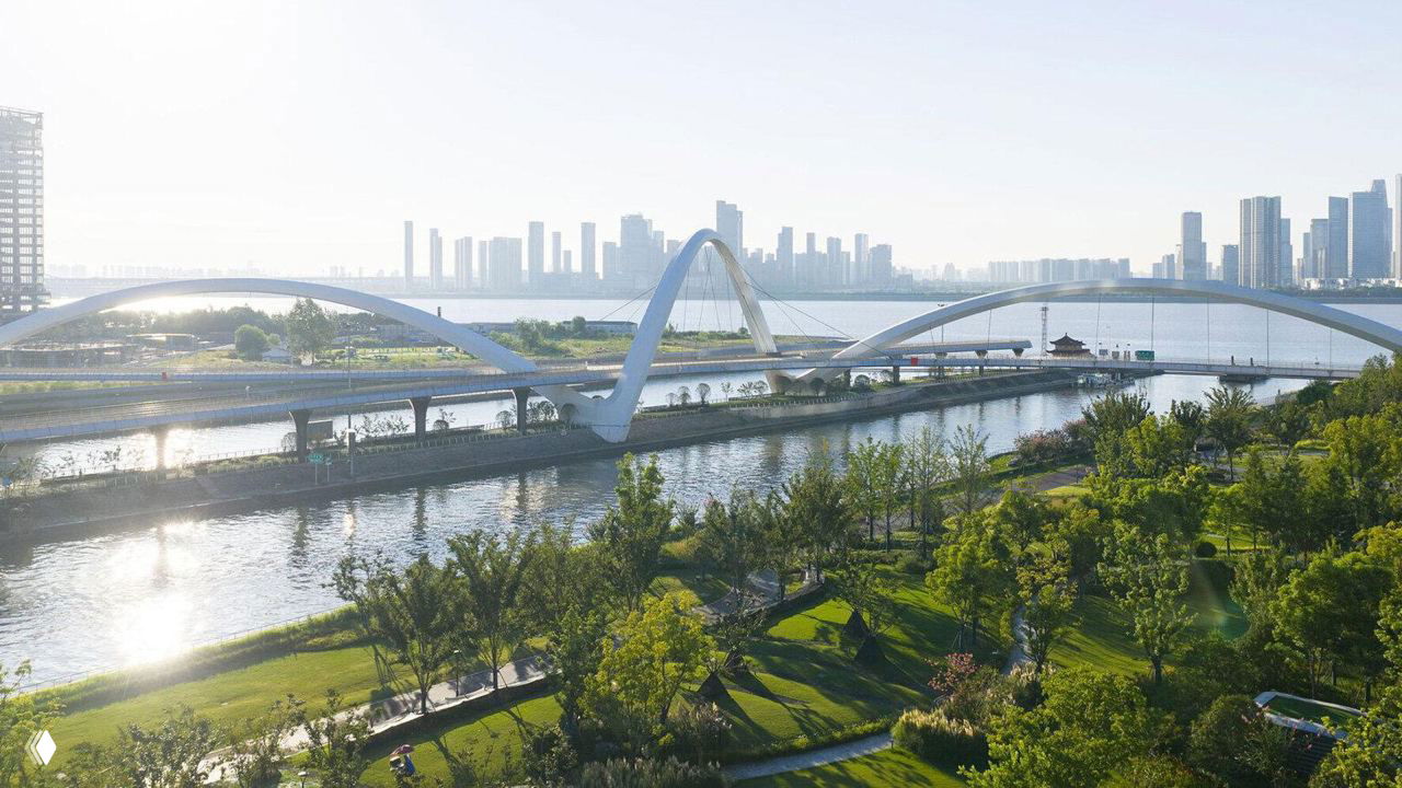 Аэрофотография белого пешеходно‑велосипедного моста в Ханчжоу с изогнутыми стальными арками над Великим каналом и зелёными набережными.