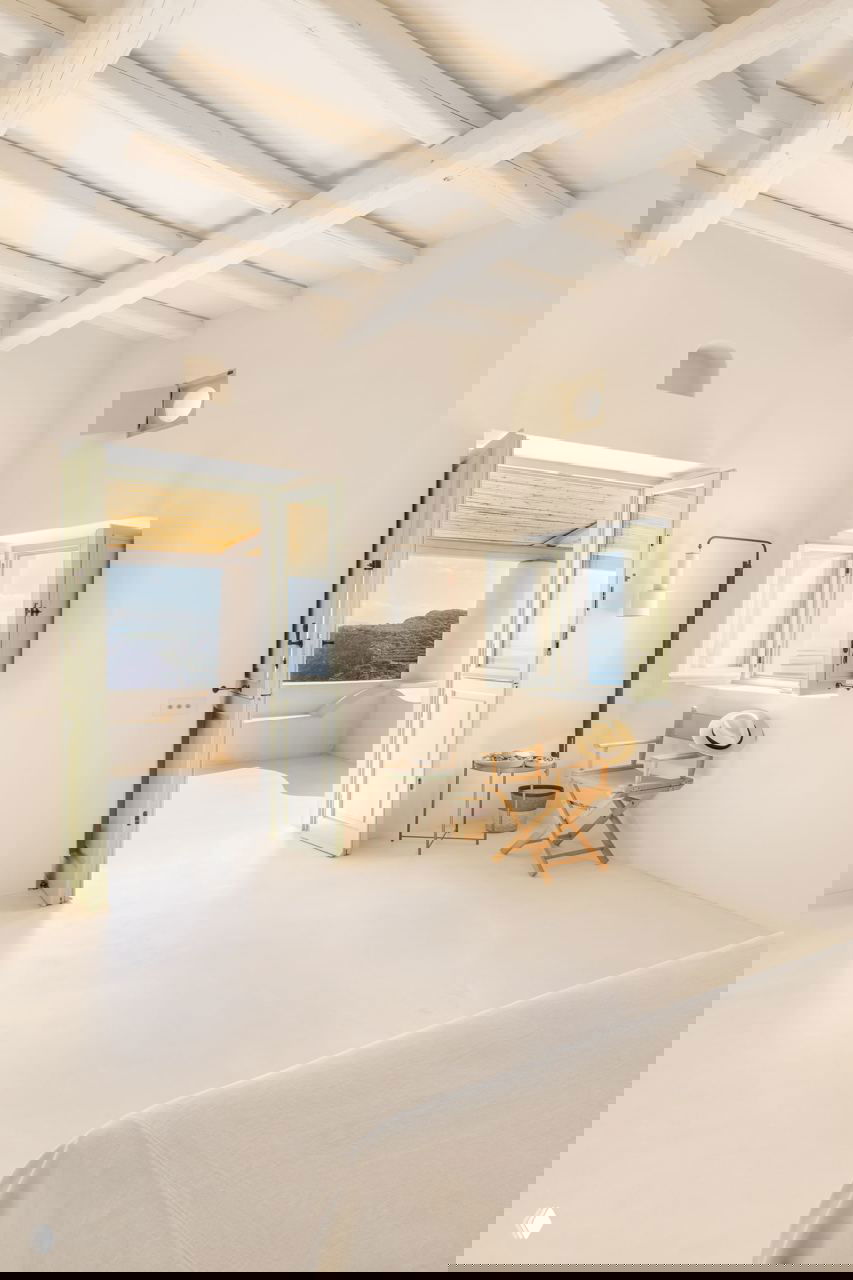 Спальня с открытыми дверями на террасу, светлые потолочные балки, деревянный складной стул и аксессуары, создающие образ отдыха и простоты.