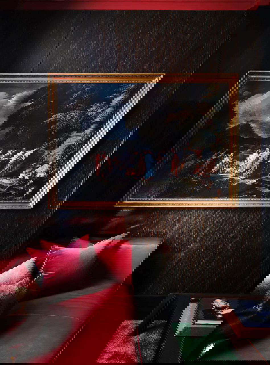 Тёмная уютная зона с малиновым кожаным диваном на фоне стен из темного дерева и большим классическим живописным полотном в раме.