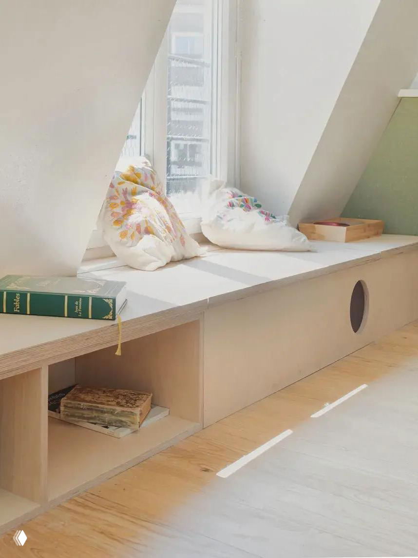 Светлый подоконник-лавка в мансарде с мягкими подушками и книжными полками под сиденьем, солнечный дневной свет в интерьере.