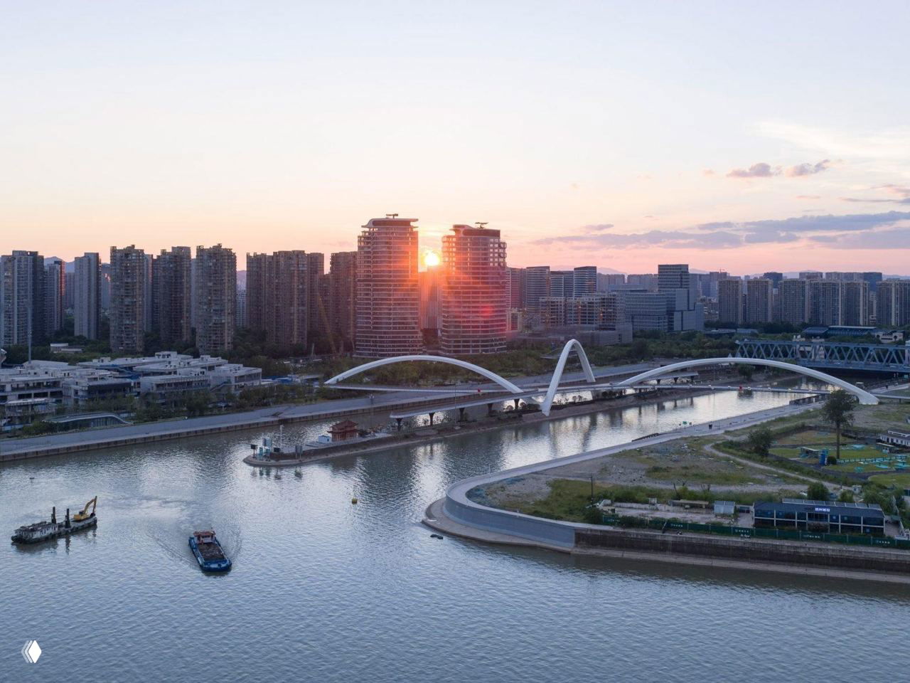 Вид на мост Zaha Hadid на закате: белые арки пересекают канал, на горизонте плотная городская застройка и отражения в воде.