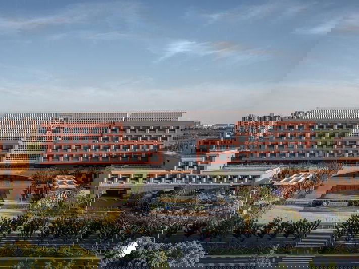 Фасад главного корпуса кампуса: строгая геометрия, красновато-коричневые керамические панели и симметричная композиция на фоне городского окружения.