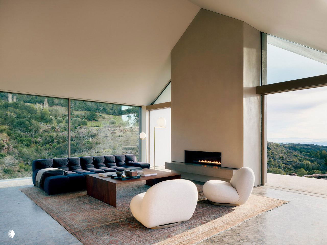 Гостиная с панорамными окнами и камином: низкий модульный диван, округлые кресла и журнальный стол на фоне широкого вида на холмы.