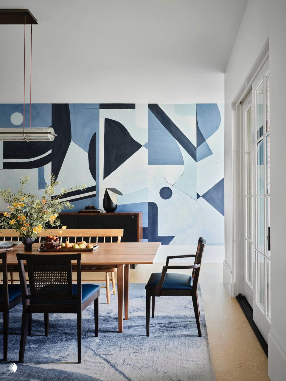 Современная столовая с геометрической абстрактной росписью на одной стене в голубо-синих тонах, деревянный стол и стулья, большое окно справа.