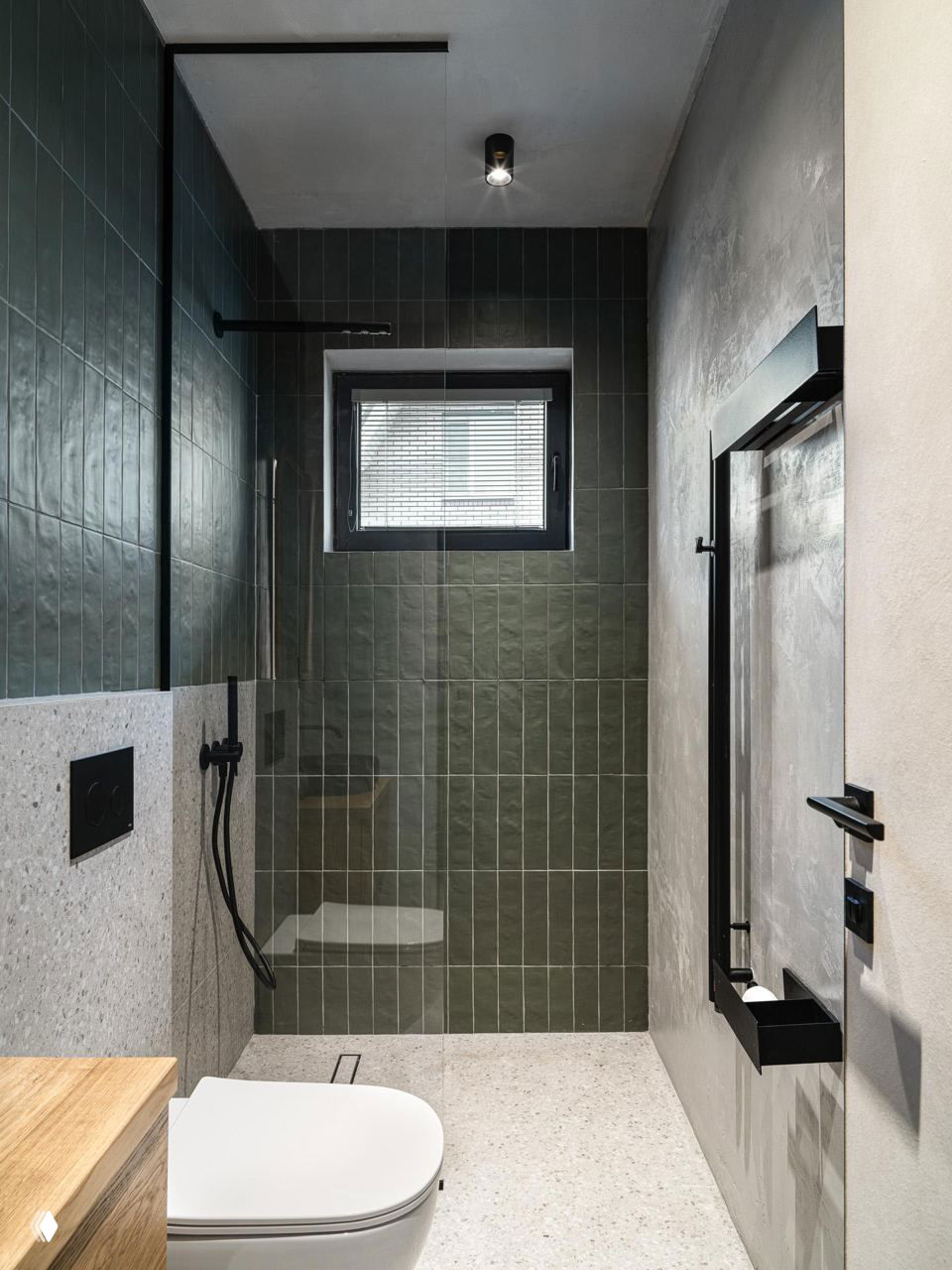 Современный душевой уголок с тёмно-зелёной вертикальной плиткой, стеклянной перегородкой, матовой фурнитурой и светлой терраццо-полкой в санузле.