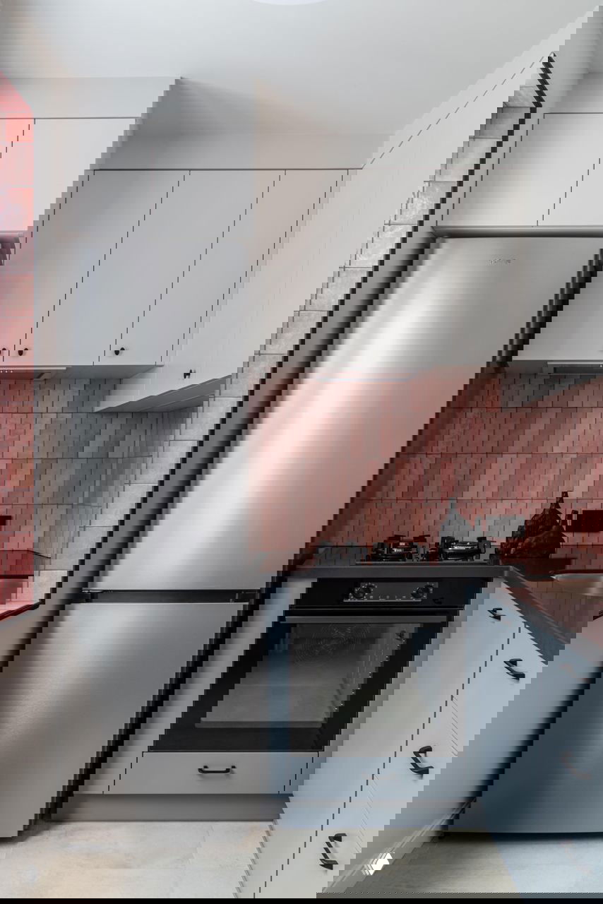 Мини-кухня с розовой плиткой фартука, встроенным духовым шкафом и серебристым холодильником; светлая мебель и тёмная столешница в небольшой планировке.