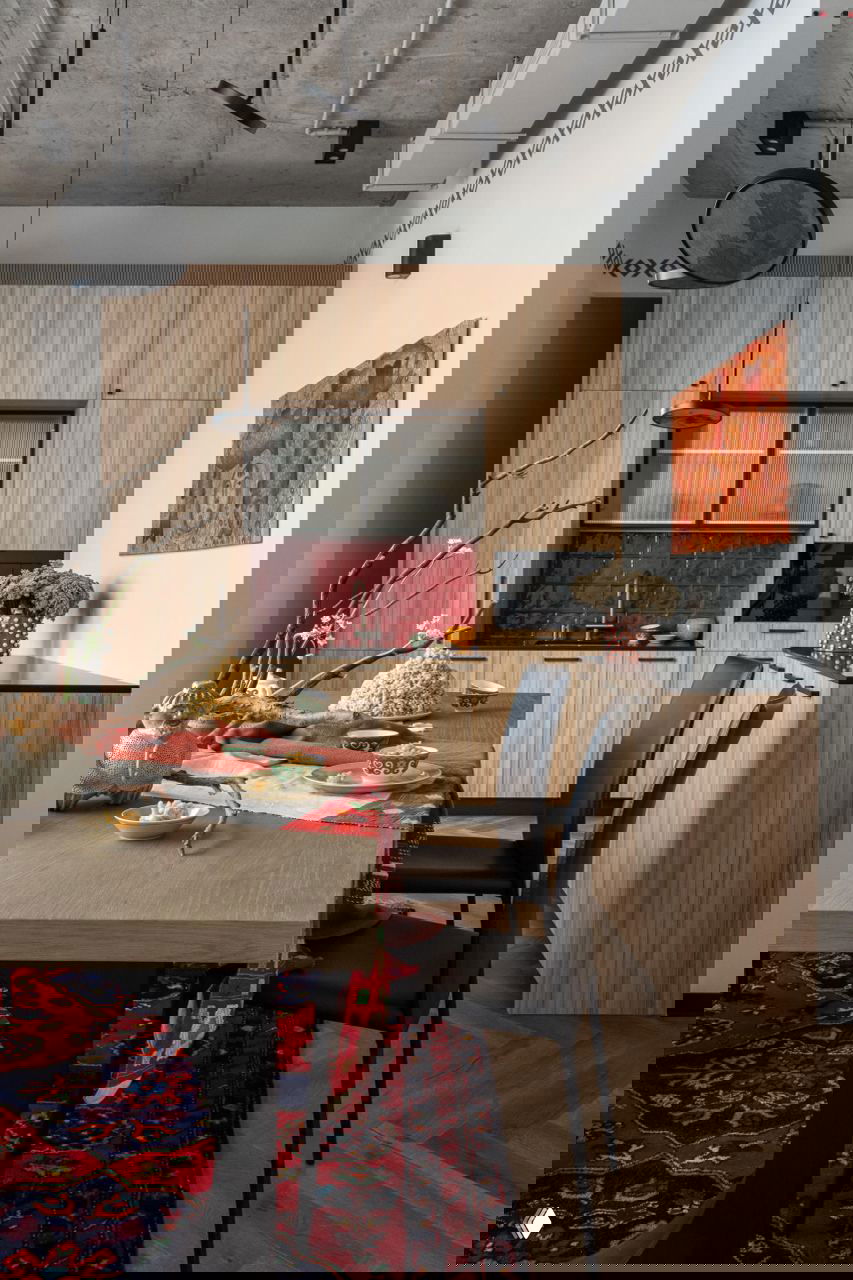 Кухонная зона в офисе: деревянные фасады, красная плитка фартука, стол с декоративной тканью и посудой в этническом стиле на фоне.