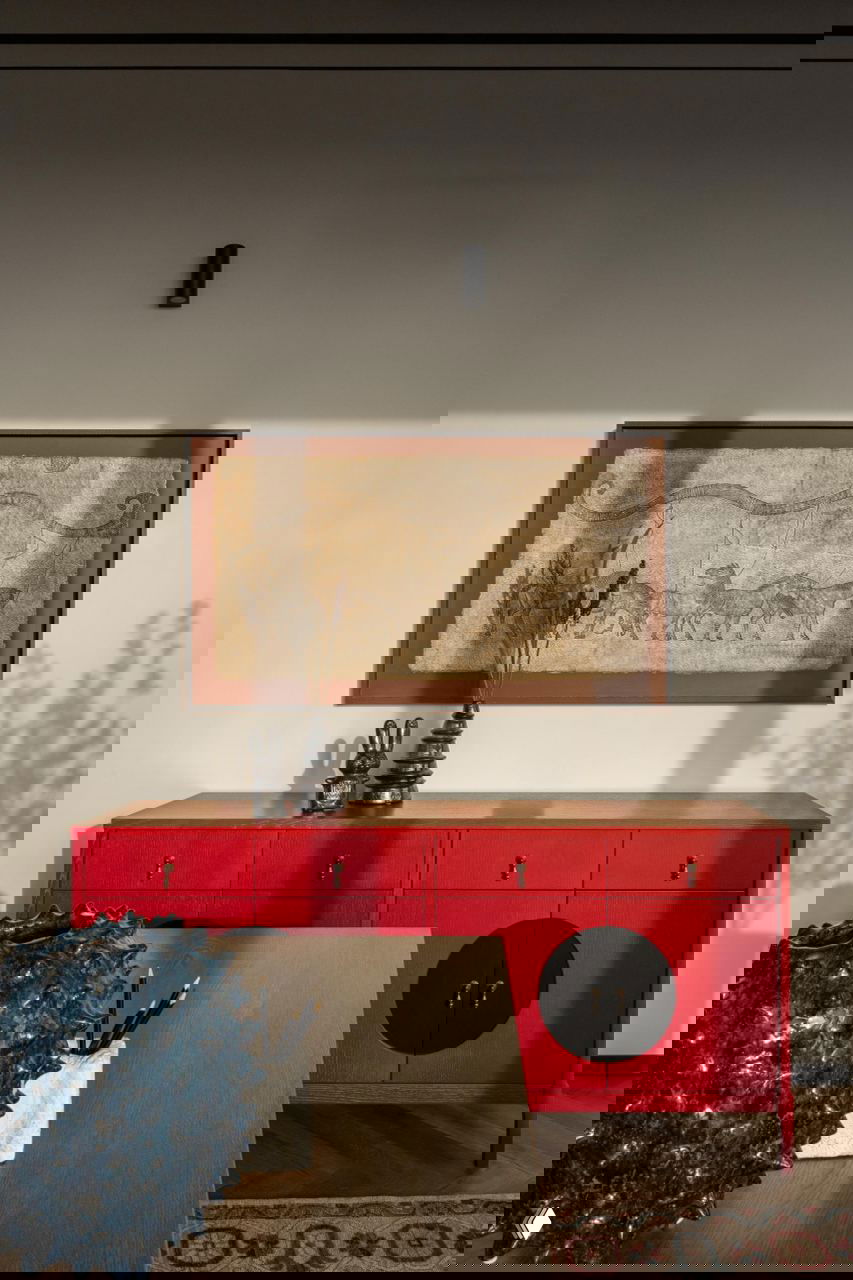 Красный комод и настенная работа: светлая стена с картиной в рамке, на переднем плане декоративные предметы и части стола.