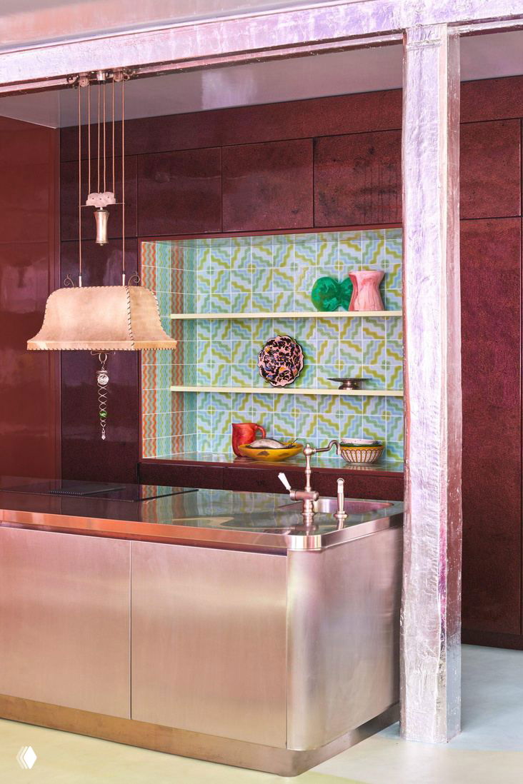 Интерьер кухни с металлическим островом, глянцевыми стенами и мозаичной нишей — яркая палитра и отражающие поверхности в стиле Uchronia в Париже.