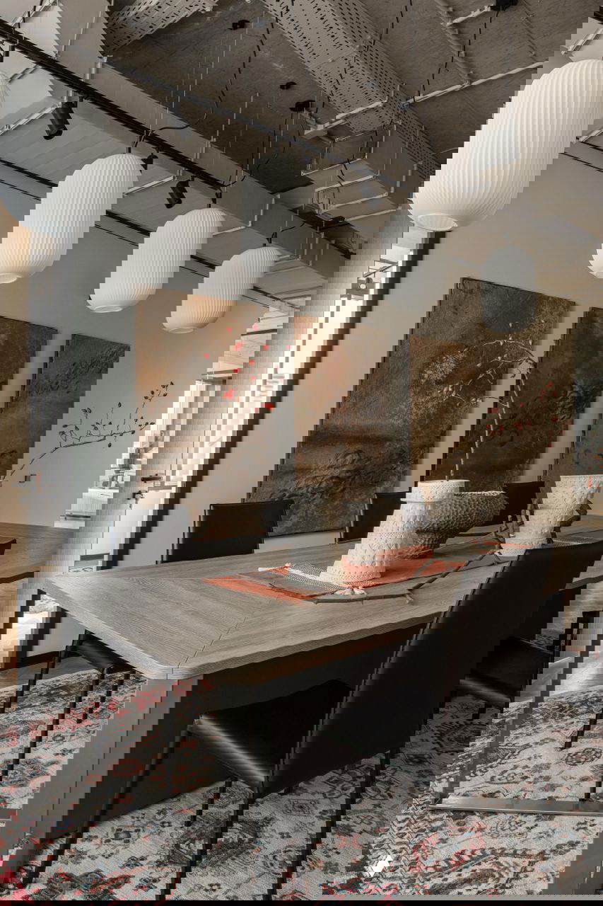 Столовая зона с тремя большими панно на стене, длинный стол, вазы и подвесные белые фонари на фоне этнического ковра под столом.