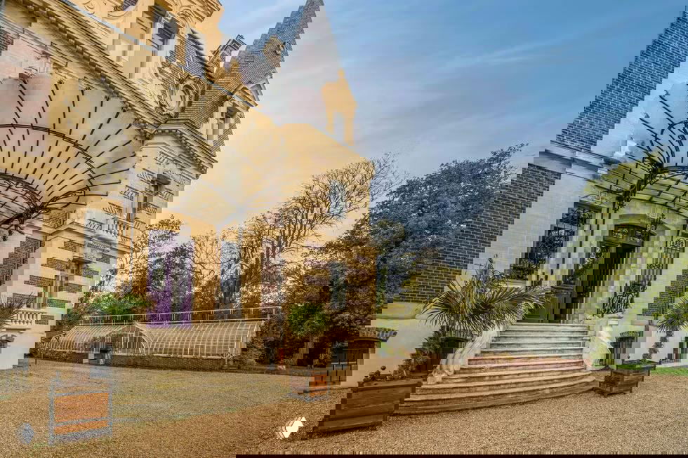 Фасад неоготического особняка Жозефины Бейкер рядом с Парижем: крыльцо с кованым навесом, пара пальм в кадре и вид на стеклянную оранжерею на участке.