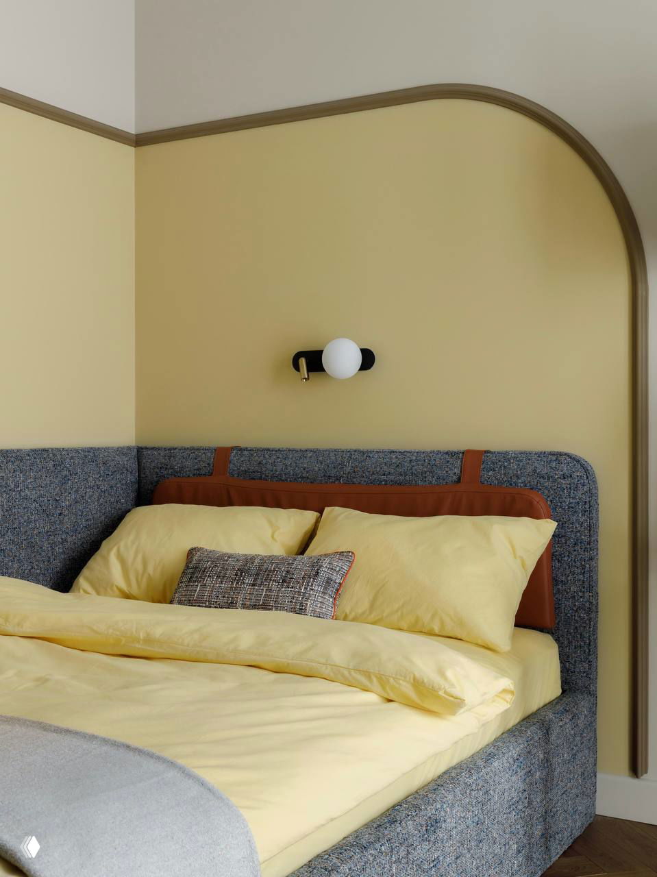 Спальня с нежно-жёлтой акцентной стеной и арочным декоративным элементом, изголовье кровати и тёплые постельные оттенки создают мягкий атмосферный интерьер.